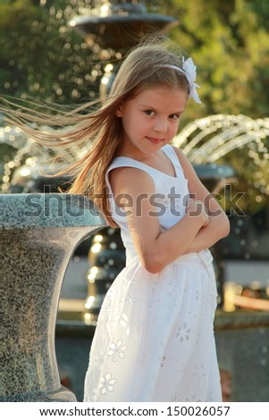 Lovely little girl in white dress sitting on parapet near the fountain in the summer hottest day/Portrait of beautiful girl near the fountain
