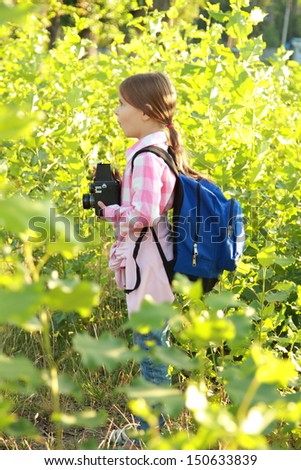 Cute young girl with a school backpack and a camera takes pictures of plants for a school lesson in the forest