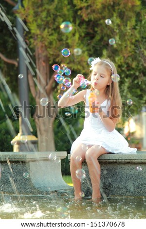 Beautiful smiling girl in a white dress flounder feet in the fountain and blowing soap bubbles in summer park/Little girl having fun playing and enjoying the spray of the fountain on a hot day