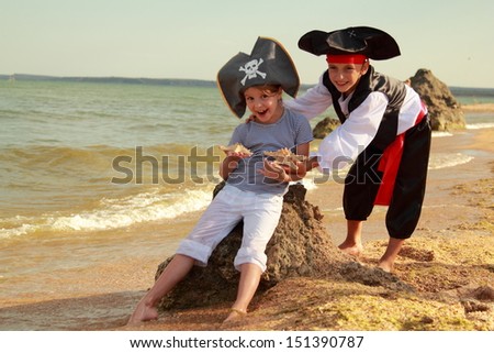 Cute little boy in a pirate costume and a little girl in a hat with a skeleton symbol of piracy are sitting on a large rock on the beach on a summer day