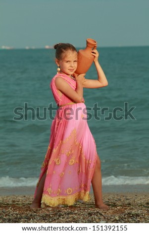 Adorable little girl with a sweet smile dressed in antique style holds an amphora on the sea/Greek antique style