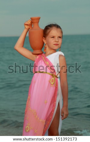 Smiling cute little girl in a beautiful dress in Greek style with an amphora stands barefoot in the sea water at the seaside