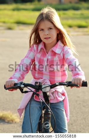 Charming young girl in blue jeans rides a bicycle in a park on a sunny day/Caucasian smiling child playing sports on a bicycle in a park outdoors
