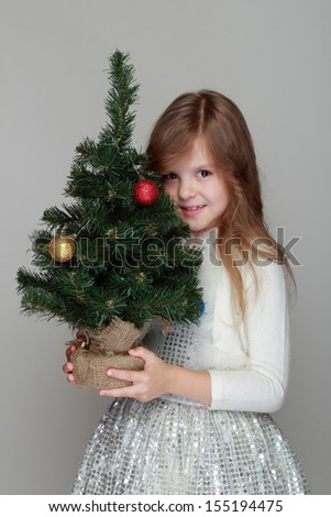 cheerful little girl in a shiny dress holding a small Christmas tree with balls on a gray background for the New Year