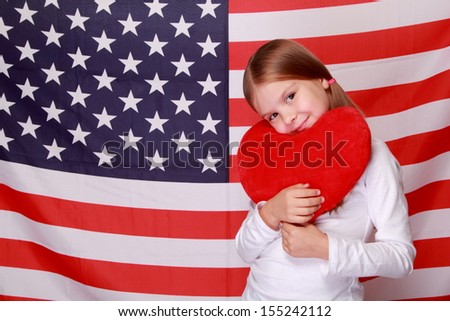 Image of a beautiful cheerful little girl with a sweet smile, holding a big red heart on the background of the American flag