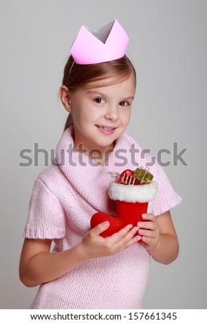 Portrait of charming smiling child in a knitted dress with a crown on his head holding Christmas boots with gifts on gray background