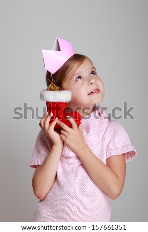 Adorable little girl in a knitted dress with a crown on his head holding Christmas boots on a gray background