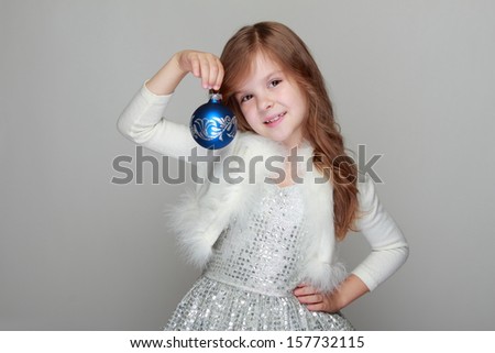 Studio image of a charming young girl in a shiny dress with fur cape holding a Christmas decoration on Holiday