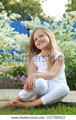 Portrait of a cute girl with long healthy hair in summer clothes walks in the yard on a background of green trees