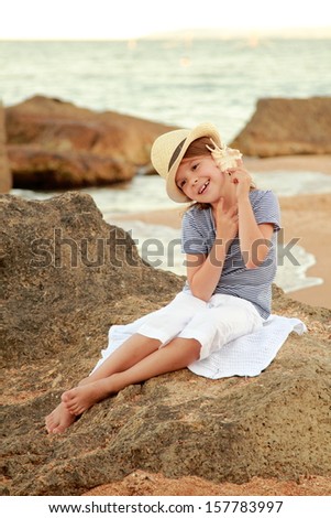 Cute joyful little girl in a summer hat sits on the shore of the blue sea on a summer theme