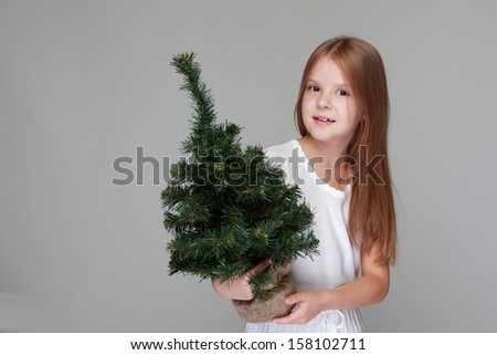 Caucasian happy little girl with a beautiful smile holding a small Christmas tree to decorate the new year on a gray background