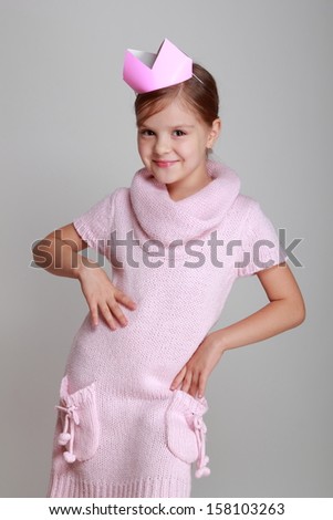 Portrait of charming smiling child in a pink knitted dress with a pink crown on his head on a gray background