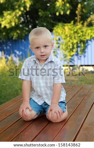 Cheerful cute little blond boy climbed up on a wooden table and laughing outdoors/Child climbed up with his feet on the table