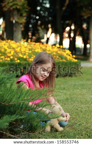 Cute little happy girl hugging brown teddy bear and sitting down on green grass meadow