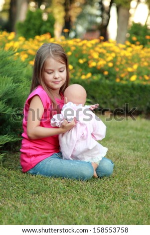 Charming cheerful little girl with long hair playing with a doll in a summer park on the green grass near the flower beds