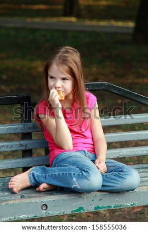Cute little girl in a red shirt eating a pie on a park bench