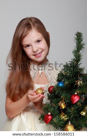 Portrait of a cute child in a knitted dress decorates a Christmas tree on a gray background on Holiday/Smiling girl near a Christmas tree