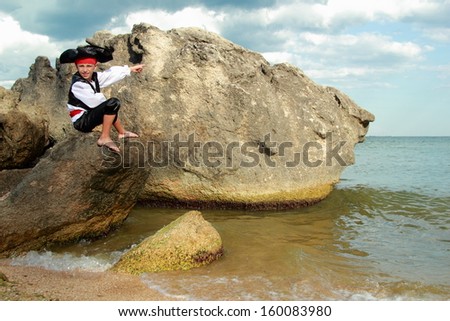 Angry little boy dressed as a pirate is sitting on a large rock in the sea and waiting for the ship