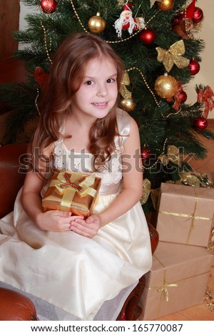 Adorable smiley princess wearing white dress and posing on camera with nice gift box over Christmas tree/Pretty little girl with Christmas gift on Holiday theme