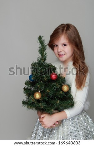 Charming cheerful little girl in a shiny dress holding a small Christmas tree with balls on a gray background for the New Year