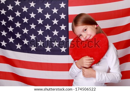 Image of a beautiful cheerful little girl with a sweet smile, holding a big red heart on the background of the American flag on Holiday