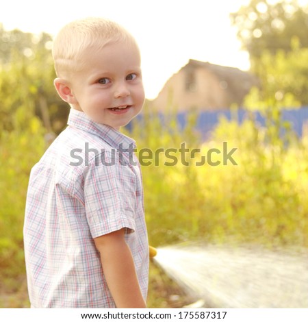Cute little blond boy in summer clothes watering the lawn with a hose in a hot summer day