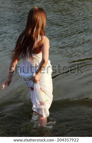Joyful cute little girl in a white dress with long hair playing in the water at the lake in the evening