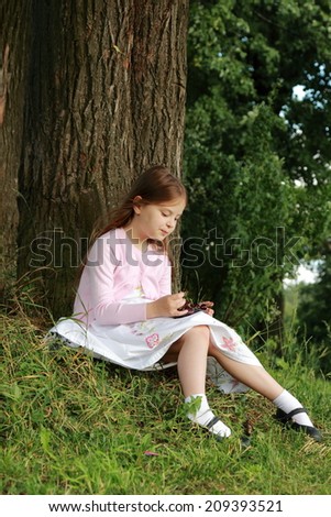 Adorable happy little girl in white dress sitting under a tree and eats cherries on a sunny summer day in the woods
