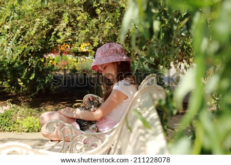 Cute happy little girl with a kitten sitting on a wooden bench in the sunny summer garden outdoor on a healthy holiday