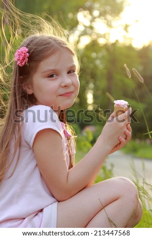 Cute young girl with a sweet smile in white dress sitting on grass and eating ice cream on a hot sunny day