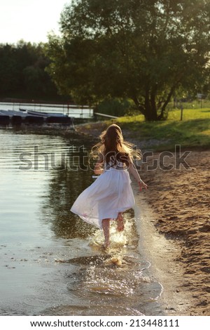 Emotional cheerful little girl in a white dress runs along the lake shore in summer