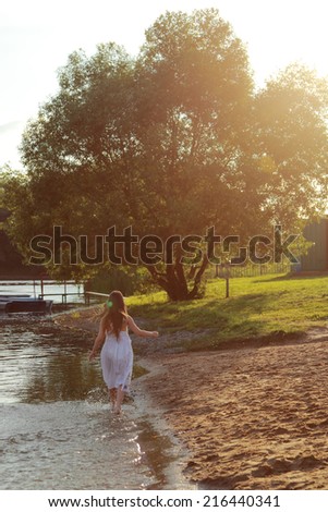 Beautiful happy young girl in a white dress running barefoot through the water in the sun at sunset