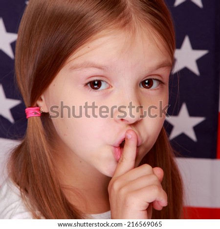 Happy smiling little girl put her finger to her lips as a sign of silence on the background of a large U.S. flag