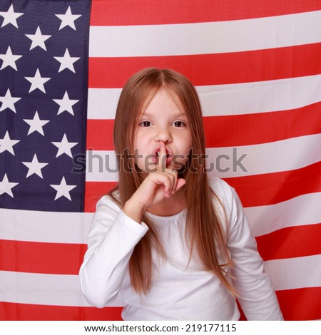 Happy smiling little girl put her finger to her lips as a sign of silence on the background of the American flag