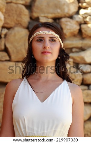 Attractive young woman in a long white wedding dress with beautiful hairstyle on background of an old stone wall as the ancient Greek goddess