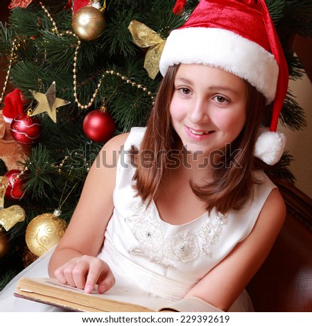 little miss santa sitting over the christmas tree and reading vintage big book on Holiday theme/Beautiful little girl wearing red and white santa\'s hat and holding old fashion book on Vintage theme