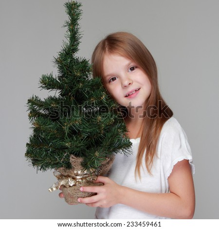 Caucasian happy little girl with a beautiful smile holding a small Christmas tree to decorate the new year on a gray background