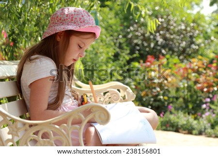Cute smiling little girl with enthusiasm draws in album sitting in the summer park bench