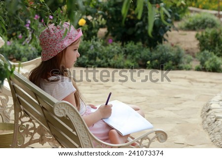 Charming concentrated little girl draws on the album to draw sitting in the summer park outdoors on bench