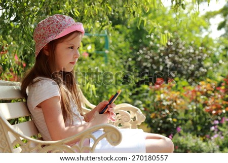 Charming concentrated little girl draws on the album to draw sitting in the summer park outdoors on bench