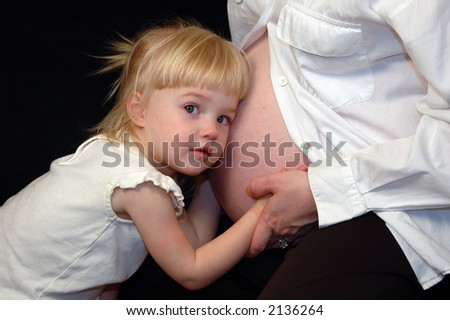 Pregnant mother and daughter embracing over black background.