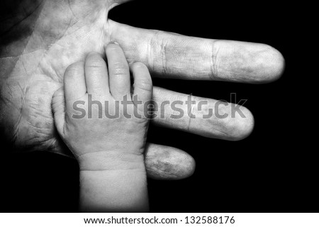 A black and white close up of a soft newborn baby\'s hand contrasted by his or her Father\'s gruff weathered hand