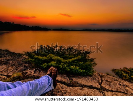An orange sunset over a calm lake.  A mans crossed legs and feet shown outstretched relaxing on a rocky shoreline looking over the lake.  Picture is showing the mans perspective.