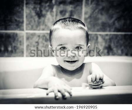 A bright eyed happy young boy sits in the bathtub looking out holding a toy fish.   Black and white image toned for a vintage looked.