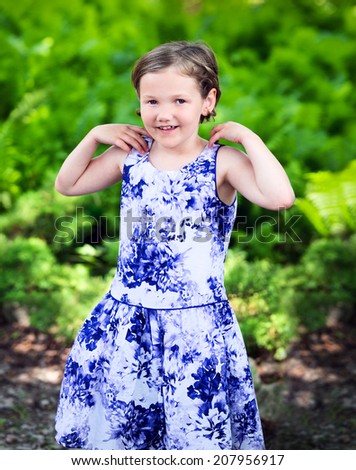 A portrait of a little girl smiling in a summer dress standing outside in a garden or park setting.