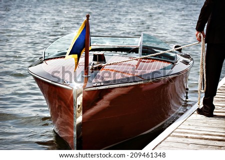 A docked vintage wooden motor boat.  A man stands on the dock holding the mooring line.
