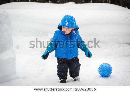 A little boy outside in the snow in the winter time with a ball on the ground next to him.