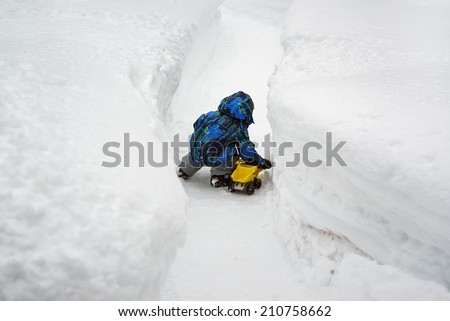 A little boy dressed in a snowsuit is crouched down outside playing with a yellow toy dump truck in a cleared snow path and there are deep snow banks on either side of him.