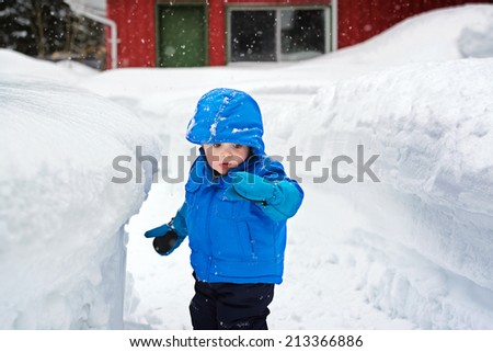 On a snowy day a little boy is standing next to a deep snow bank which is as tall as he is.   He is looking at snow on his glove.