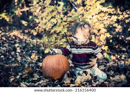A boy sits beside a pumpkin outside on the leaf covered ground in a yard or park holding on to the stem during the autumn season.  Filtered to give vintage, faded look.